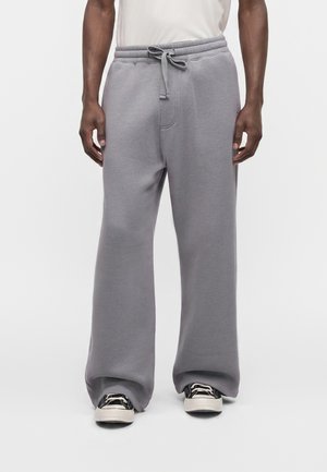 Homme portant un pantalon de survêtement gris ample à taille à cordon et des baskets noires et blanches, debout sur fond blanc.