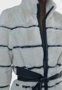 Bílý kožich s černými akcenty, s prošívaným vzorem, vysokým límcem, plným zipem a koženým páskem u pasu.