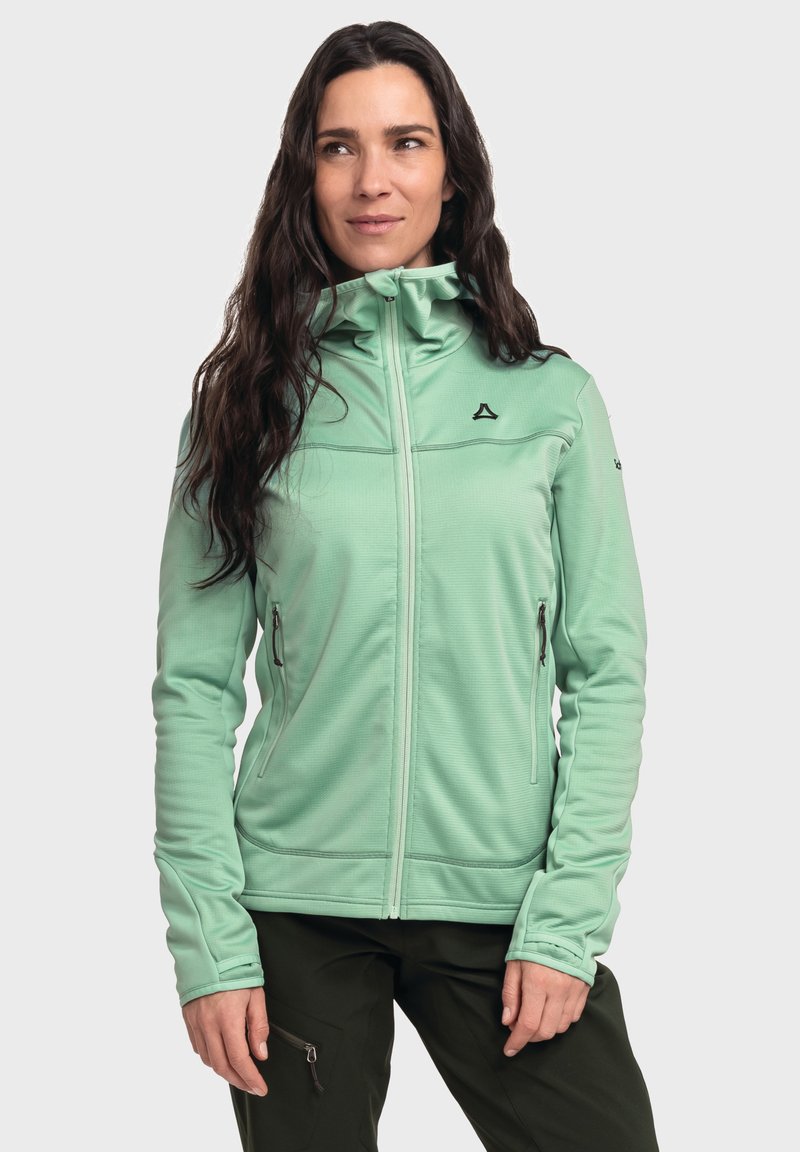 Helles grünes Zip-Jackett mit hohem Kragen, strukturiertem Stoff, frontalen Reißverschlusstaschen und einem schwarzen Logo auf der Brust. Lange Ärmel mit Daumenlöchern.