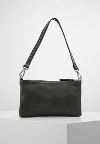 Pochette en cuir noir avec clous argentés sur la bandoulière amovible, fermeture éclair supérieure et logo discret sur le devant, présentée sur une surface blanche.