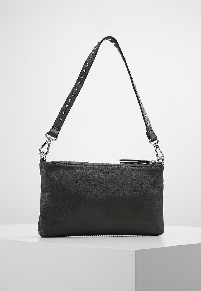 Pochette en cuir noir avec clous argentés sur la bandoulière amovible, fermeture éclair supérieure et logo discret sur le devant, présentée sur une surface blanche.