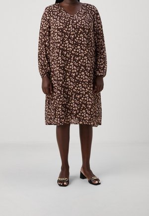 Frau trägt ein braunes, knielanges Kleid mit Blumenmuster, langen Ärmeln und schwarzen Sandalen mit Absatz und goldener Kettendetail.