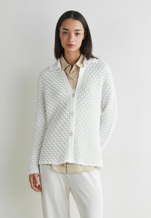 Junge Frau trägt eine strukturierte weiße, geknöpfte Strickjacke über einem beigen Hemd und weißen Hosen, steht vor einer schlichten hellen Wand.