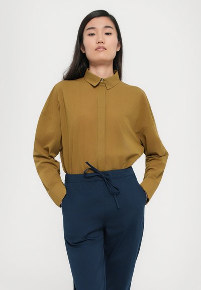 Rjava srajca s kratkimi rokavi in ovratnikom, v kombinaciji z modrimi hlačami na vrvico. Gladka tkanina in sproščen kroj.