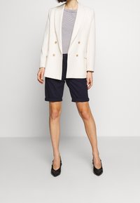 Dvouřadý krémový blazer se zlatými knoflíky, spárovaný s námořnickými krátkými kalhotami a špičatými černými lodičkami, pod kterým je pruhované tričko.