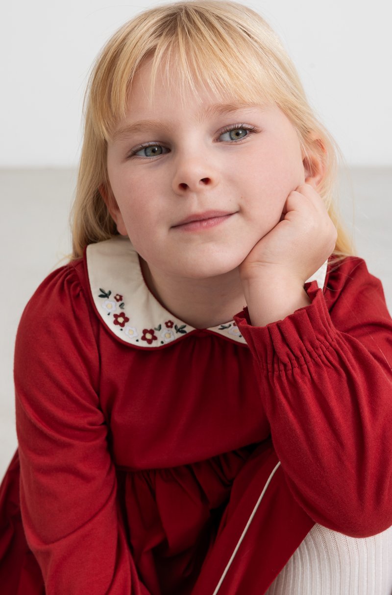 Abito rosso con maniche lunghe, vita arricciata e colletto bianco con ricamo floreale. Tessuto morbido con una leggera lucentezza; accento a coste sottile sulle maniche.