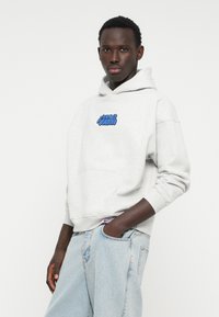 Sudadera de algodón gris con un bolsillo frontal, con letras gráficas azules en el pecho. Combinada con vaqueros de mezclilla sueltos de color azul claro.