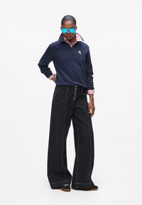 Marinblå långärmad polo med hög krage, prydd med en liten logotyp, matchad med vida mörk denimbyxor med synliga sömmar. Blå solglasögon fulländade looken.