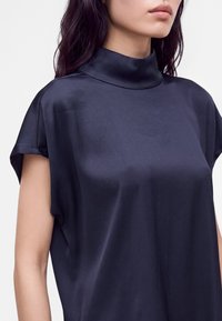 Femme portant un chemisier sans manches en satin bleu foncé à col montant, montrant le haut du torse et les lèvres sur un fond uni.