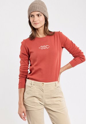 Vrouw in een beige gebreide beanie, roestkleurig shirt met lange mouwen met een wit visontwerp, en beige broek, staand met één hand op de heup tegen een witte achtergrond.