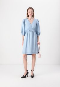 Rochie din denim albastru deschis, cu decolteu în V, mâneci bufante și talie strânsă, care se termină deasupra genunchiului. Purtați cu tocuri stiletto negre și o geantă roz.