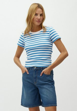 Mujer que lleva una camisa de rayas azul y blanca de manga corta metida en unos pantalones cortos de mezclilla sueltos, con las manos en los bolsillos, de pie frente a un fondo liso.