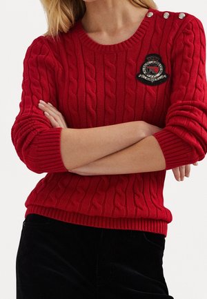 Femme bras croisés portant un pull rouge en maille torsadée avec des boutons sur l'épaule et un écusson brodé noir.