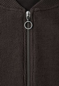 Nahaufnahme eines schwarzen, strukturierten Stoffes mit einem silbernen Metallreißverschluss, der in der Mitte einen runden Zuggriff hat.