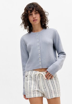 Lichtblauwe cropped sweater met lange mouwen, ronde halslijn en zeven knoopsluitingen. Gecombineerd met witte shorts met blauwe strepen.
