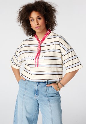 Vrouw met krullend haar draagt een gestreept oversized overhemd, rode sjaal om de nek, lichtblauwe high-waisted jeans en armbanden, met haar handen in de zakken.