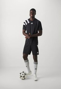 Fekete adidas sportfelszerelés fehér csíkokkal, rövid ujjú inggel és rövidnadrággal. A modell egy focilabdával áll, térdig érő zoknit visel.