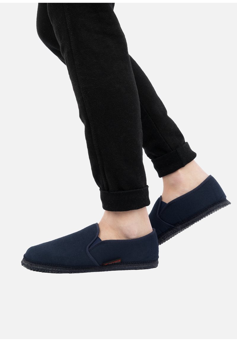 Dark navy slip-on shoes made of soft fabric with a flexible sole and elastic side accents. Worn with black tapered sweatpants.