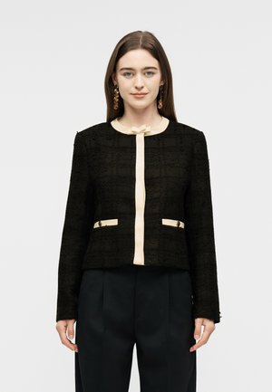 Mladá žena otočená čelom k nám, oblečená v čiernej veste s textúrou, krémovým lemom a ozdobnou mašľou, v kombinácii s čiernymi nohavicami a visiace náušnice.