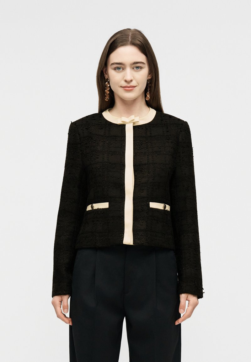 Jeune femme de face portant une veste noire texturée avec des bordures crème et un nœud décoratif, associée à un pantalon noir et des boucles d'oreilles pendantes.