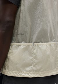 Oldalnézet egy személyről, aki világos bézs színű, texturált dzsekit visel, zsebekkel és a szöveten részben látható "cispiritual" felirattal.