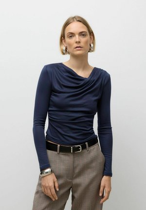 Navy long-sleeve top with draped neckline and fitted design, paired with plaid trousers and a black belt. Metallic bangle on wrist.