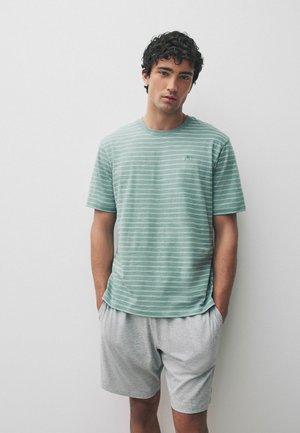 Junger Mann mit lockigem dunklem Haar, der ein grün-weiß gestreiftes T-Shirt und hellgraue Shorts trägt und mit den Händen in den Taschen steht.