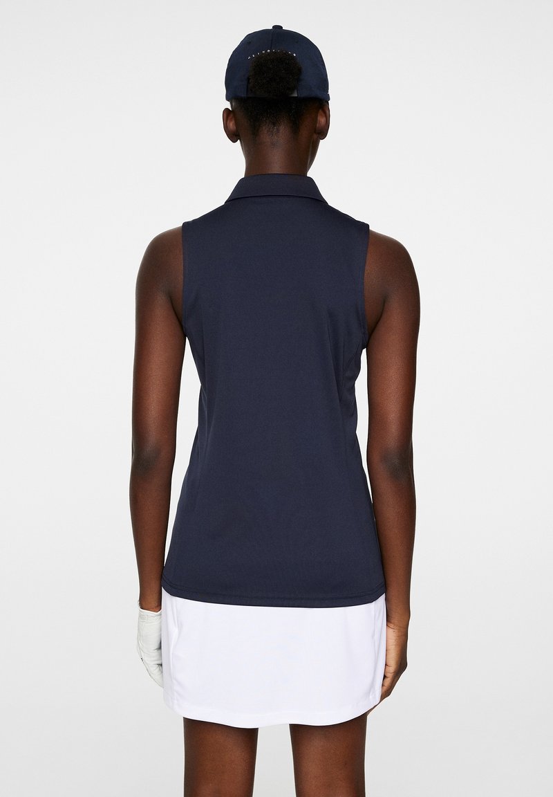 Polo senza maniche blu navy con una texture morbida, abbinato a una gonna bianca, con colletto e una vestibilità aderente. Guanto bianco visibile sulla mano.