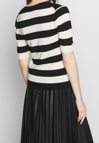 Žena v černobílém vodorovně pruhovaném svetru s krátkým rukávem a černé plisé sukni, zobrazená zezadu.