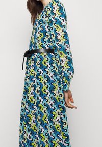 Robe à manches longues présentant un motif floral multicolore avec des fleurs bleues, vertes, noires et blanches ; cintrée avec une ceinture noire à la taille.