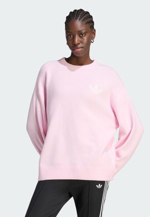 Vrouw met gevlochten haar, gekleed in een lichtroze trui en zwarte Adidas-broek met witte strepen, staand tegen een effen lichte achtergrond.