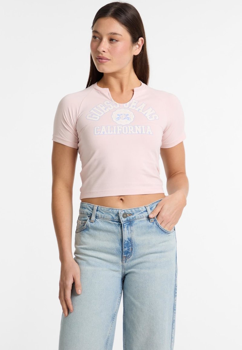 Camiseta rosa corta con cuello redondo, mangas cortas y logo de "Guess Jeans California" en azul claro. Se combina con jeans azul claro.