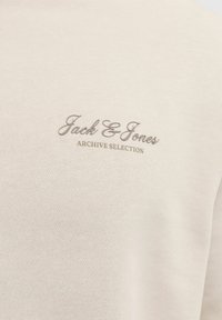 Svetlo bež majica z teksturirano površino, z vdelanim temno sivim logotipom "Jack & Jones" in napisom "ARCHIVE SELECTION" spodaj.