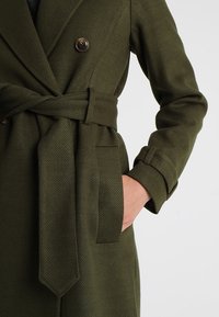 Olivgrüner Wollmix-Mantel mit doppelter Knopfleiste, Reverskragen, Taillengürtel und seitlichen Taschen, ausgestattet mit einem Fischgrätmuster.