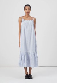 Modrá a biela pruhovaná bezrukávová šaty s nariaseným výstrihom a volánkovým lemom, vyrobené z ľahkej látky; doplnené čiernymi topánkami.