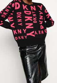 Pull noir avec un lettrage rose vif "DKNY", coupe décontractée, manches longues. Associé à une jupe noire brillante à motif crocodile.