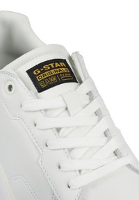 Baskets en cuir blanc avec un bout rond, lacets blancs plats et une étiquette de marque noire et dorée sur le côté. Accents texturés sur le dessus.