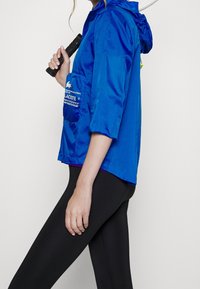 Blå, glänsande, huva jacka med en känguruficka och logotypdetalj på framsidan. Buren över svarta leggings, sidovy.