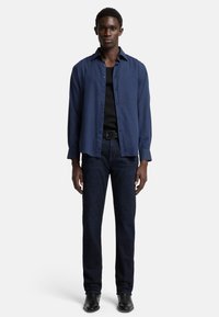 Camicia blu navy a bottone in tessuto leggero, indossata sopra una canotta nera, abbinata a jeans in denim scuro e scarpe nere.