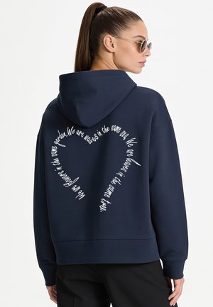 Femme aux cheveux bruns en queue de cheval, portant des lunettes de soleil rondes et un sweat à capuche bleu marine avec un texte blanc en forme de cœur dans le dos.