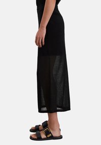 Schwarzer Maxikleid mit einem transparenten unteren Abschnitt, aus leichtem Stoff gefertigt. Kombiniert mit flachen schwarzen Sandalen, die über eine goldene Schnalle verfügen.