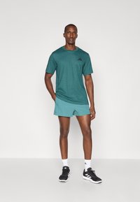 T-shirt de sport teal avec logo noir, associé à un short teal. Baskets noires avec accents blancs. Texture lisse, design décontracté.