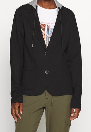 Muž ve černém saku s šedou kapucí, bílém tričku s potiskem a olivově zelených kalhotách s boční kapsou.