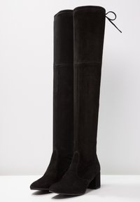 Botas negras hasta el muslo hechas de suave ante, con punta afilada, tacón cuadrado y un detalle de encaje en la parte superior. Textura suave y diseño elegante.