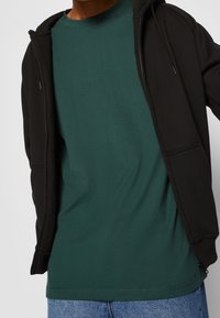 Sudadera negra con cremallera sobre una camiseta verde oscuro, con cuello redondo y mangas cortas, combinada con pantalones vaqueros azules.
