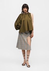 Γυναίκα φορούν ελιά πράσινο τοπ ενός ώμου με φουσκωτό μανίκι και καρό φούστα με μεγάλο άνοιγμα πάνω από το γόνατο, μαύρα σανδάλια με λουριά.