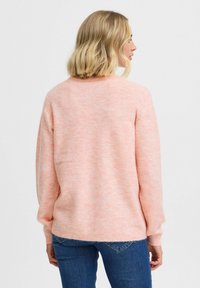Maglione rosa lavorato a maglia con una finitura morbida e testurizzata. Presenta una vestibilità comoda e polsini a coste, indossato sopra dei jeans blu.