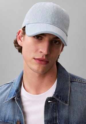 Casquette en denim bleu clair avec un motif texturé subtil, dotée d'une visière courbée. La casquette est portée avec un t-shirt blanc et une veste en denim.