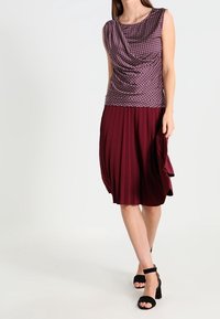 Top senza maniche di colore viola con motivo e design drappeggiato, abbinato a una gonna plissettata di un profondo bordeaux. Indossato con sandali neri con tacco alto.
