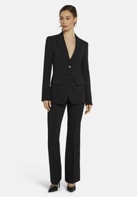 Zwarte op maat gemaakte pak met een notch lapel, twee knopen en open zakken. Slim-fit broek met een lichte flare, gecombineerd met zwarte puntige hakken.